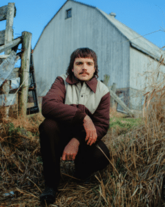 A person with a mustache squats in front of a weathered wooden barn and fence, wearing a brown and beige jacket, surrounded by dry grass under a clear blue sky.